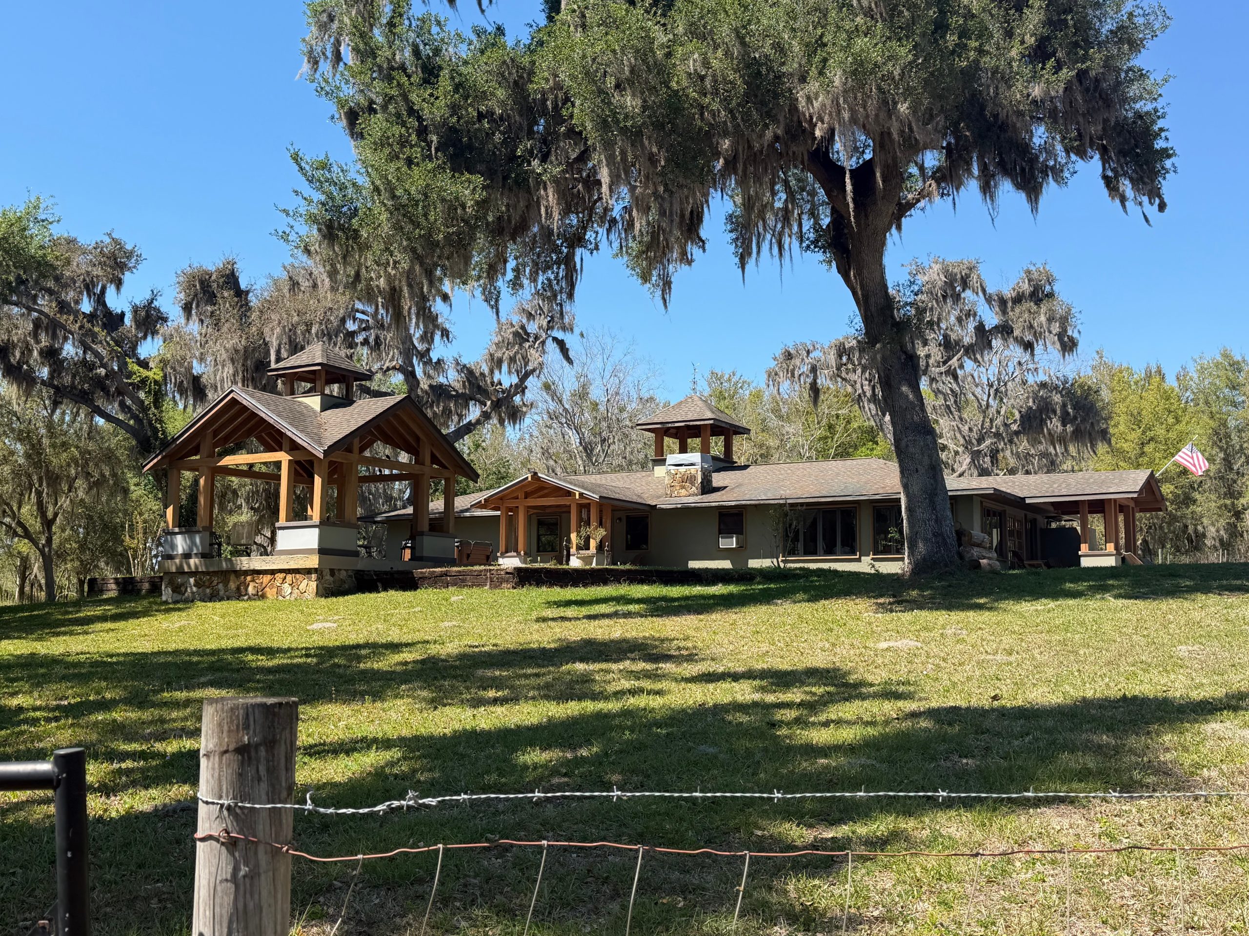 Rustic ranch house with wooden fence in a grassy field under bright daylight with trees in the background.